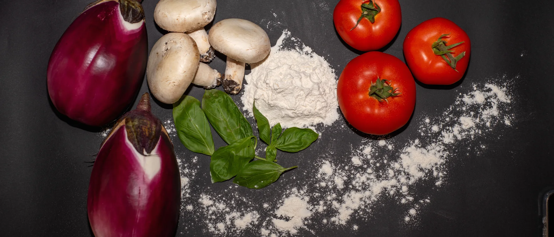 Melanzane, funghi, pomodori, basilico e farina disposti su un piano di ardesia nera